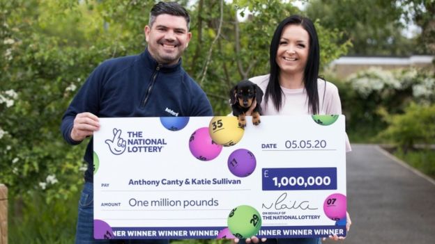 지난달 경찰관의 목숨을 구한 영국 남성 앤서니 캔티와 아내 캐티 설리번이 지난 5일(현지시간) 당첨 사실을 알리는 광고판을 든 채 포즈를 취하고 있다. 전날 밤 닥스훈트 반려견 러키 롤라를 입양했는데 다음날 아침 당첨 행운을 차지한 사실을 알게 됐다.BBC 홈페이지 캡처